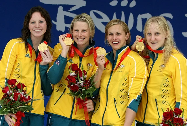 Australian women's swimming team celebrating with gold medals at the 2008 Beijing Olympics, holding flowers and smiling.