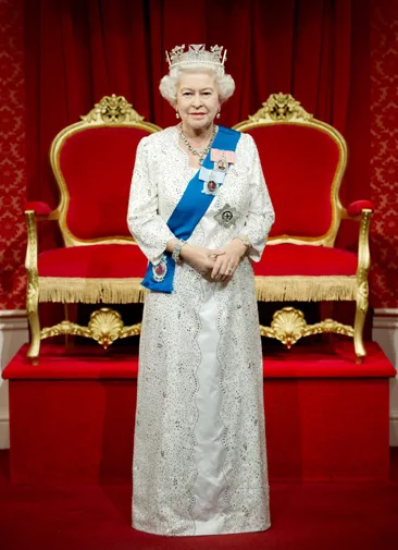 A wax figure of an elderly woman in a white gown and tiara, standing in front of a red throne with a regal pose.