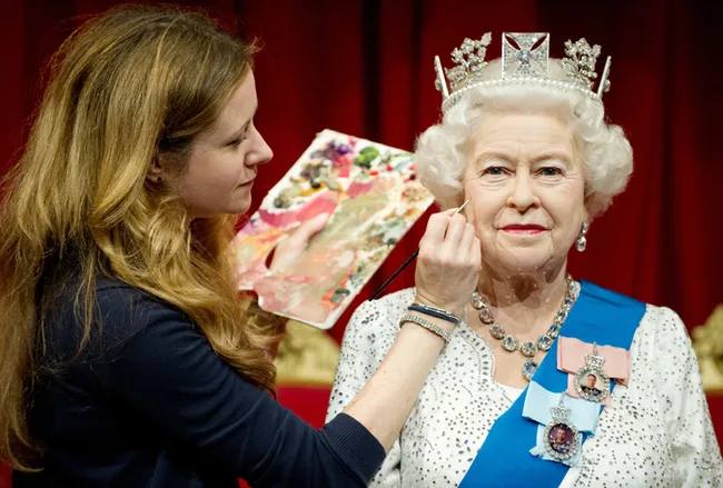 A woman touches up a wax figure of a queen, wearing a crown and blue sash, against a red background.