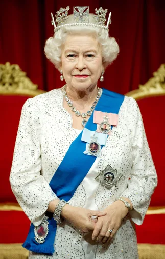 A wax figure of an elderly woman in regal attire with a crown and royal sash, standing against a red backdrop.