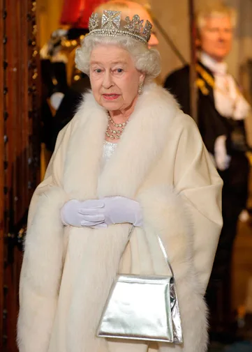 Queen Elizabeth II wearing a white fur coat, tiara, and holding a silver handbag. Background has blurred figures in formal attire.
