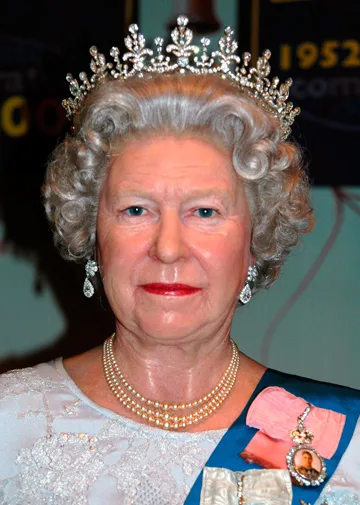 A wax figure of an elderly woman with a crown, pearl necklace, and blue sash, resembling royalty in a formal setting.