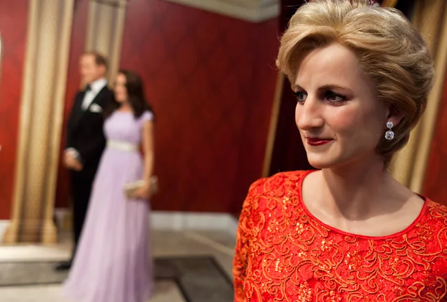 Wax figure of a woman in red dress, with two blurred figures in the background wearing formal attire.