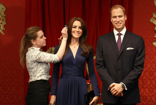 A woman adjusts the hair of a female wax figure beside a male wax figure, set against red curtains.