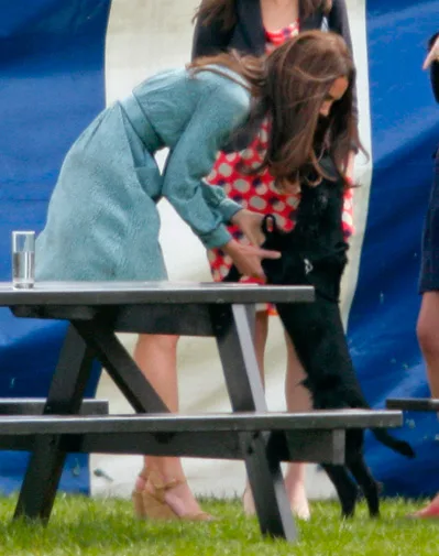 Woman in a teal dress bends to pet a black dog near a picnic table on grass with a tent in the background.