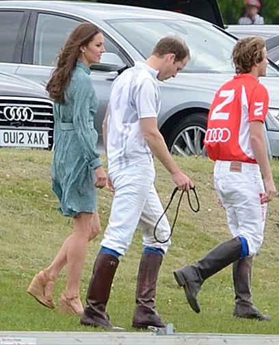 Elegant woman in green dress walks alongside two men in polo gear, one in white, one in red, near parked cars.