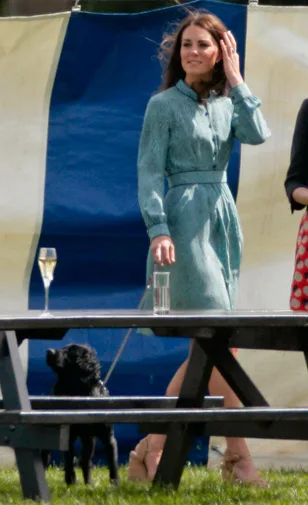 A woman in a blue dress walking a black dog past a picnic table with a glass of champagne and a glass of water.