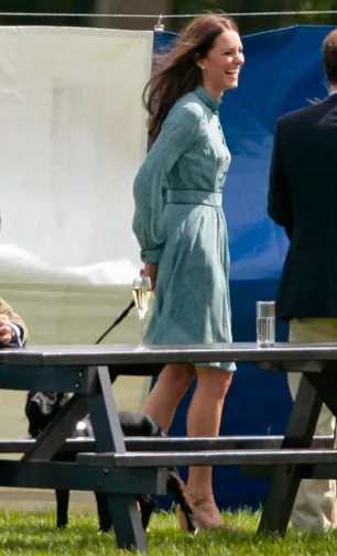 A woman in a green dress stands outdoors holding a leash and a glass, with a dog nearby and benches in the foreground.