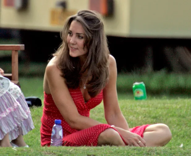 A woman in a red polka-dot dress sits on grass with a bottle beside her, looking to the side, with a relaxed expression.