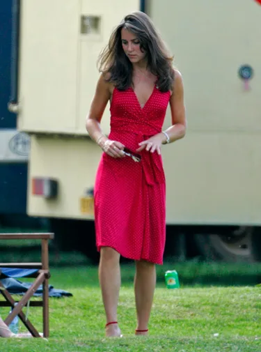 A woman in a red polka dot dress stands on grass near a chair and a trailer, holding an object in her hands.