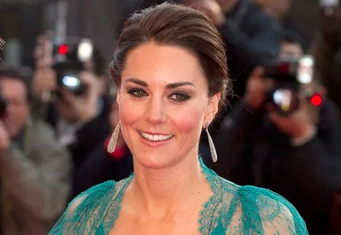 A woman smiles at a public event in a teal, sheer dress, with photographers in the background capturing the moment.
