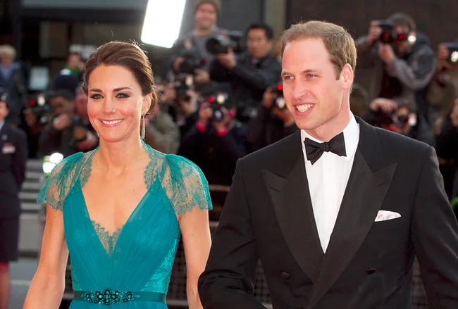 Two people in formal attire walking while being photographed by paparazzi at an event.
