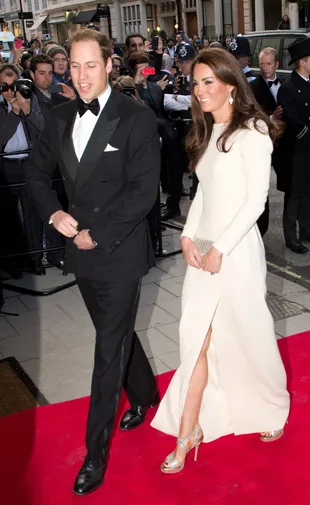 A couple in formal attire walking on a red carpet, with photographers in the background.