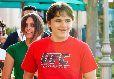 Youthful individuals smiling outdoors, one in a green shirt, another in a red UFC shirt, with greenery and a pole in the background.
