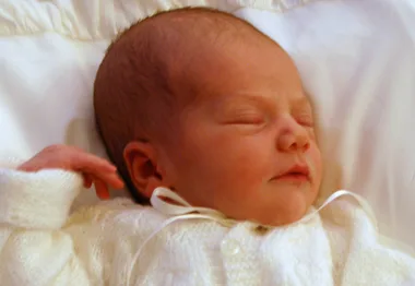 Newborn baby in a white knitted outfit peacefully asleep on a bed.