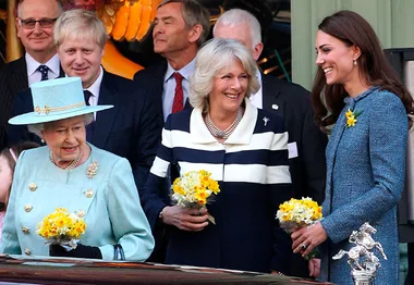 Queen, Camilla, and Kate smiling with bouquets at an outdoor event, surrounded by others in formal attire.