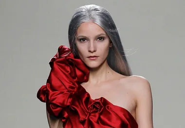 Model with long gray hair wearing an asymmetric red ruffled dress on the catwalk.