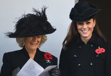 Two women in black coats and hats, smiling and wearing red poppy pins, standing together with one holding a paper.