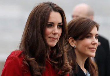 Two women stand together outdoors, one in a red coat, both smiling slightly.