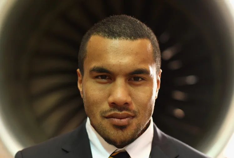 A rugby player in a suit stands in front of a turbine, looking directly at the camera.