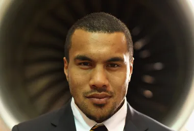 A rugby player in a suit stands in front of a turbine, looking directly at the camera.