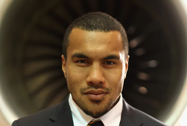 A rugby player in a suit stands in front of a turbine, looking directly at the camera.