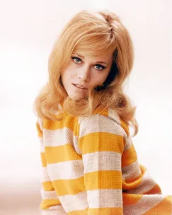 A woman with blonde hair wearing a yellow and white striped sweater, looking towards the camera with a soft focus background.