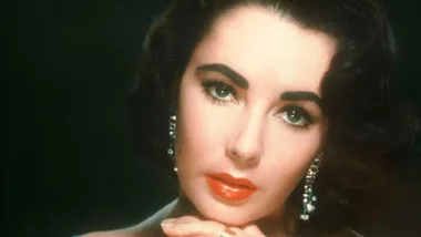 Legendary actress with striking features, wearing elegant earrings, poses with red lipstick against a dark background.