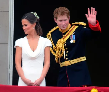 A woman in a white dress stands beside a man in a military uniform waving, on a balcony with red drapery.