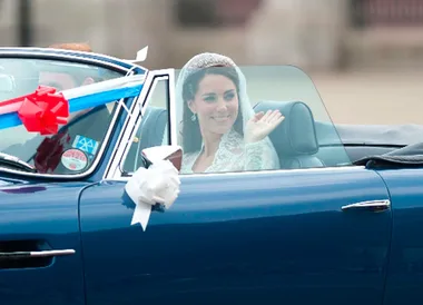 Bride in a classic blue car waves, wearing a lace dress and tiara, with wedding decorations on the vehicle.