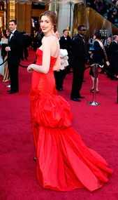 Anne Hathaway in a red strapless gown on the Oscars red carpet, smiling and turning towards the camera.