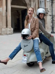 A joyful couple riding a Vespa scooter in an Italian town, both wearing helmets, with historic architecture in the background.