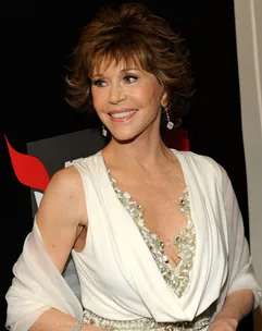 A woman in a white dress with short hair smiles, wearing dangling earrings and standing in front of a black backdrop.