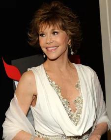 A woman in a white dress with short hair smiles, wearing dangling earrings and standing in front of a black backdrop.