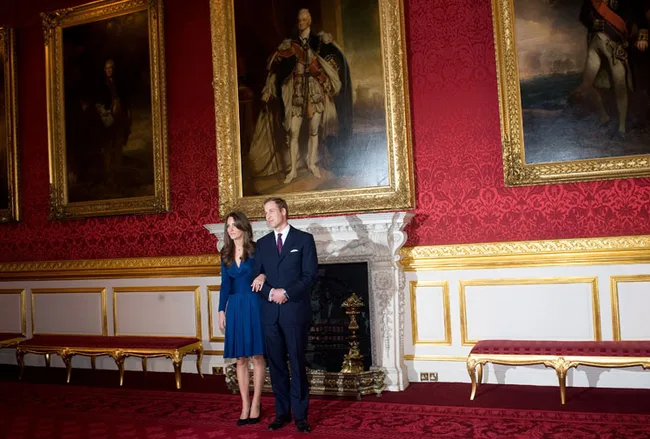 Prince William and Kate Middleton standing in a regal room with red decor and large paintings.