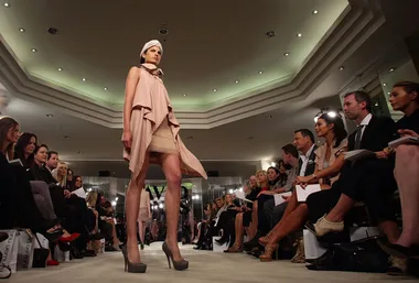 Model walking down runway in a beige layered dress with high heels, surrounded by seated audience in a well-lit venue.