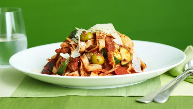 Plate of pasta with olives, zucchini, and shaved cheese on a green tablecloth, with a glass of water and cutlery nearby.