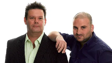 Two men posing together, one with a blazer and striped shirt, and the other in a dark shirt, against a white background.