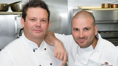 Two chefs in white uniforms pose together in a kitchen setting.
