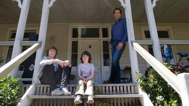 Father and two sons sit on porch steps of a house, with a bicycle in the background.