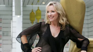 Rebecca Gibney in a sheer black blouse, smiling while leaning on a large golden chair indoors.