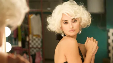 A woman with a short, curly platinum blonde wig looks at herself in a mirror, wearing elegant earrings and a black outfit.