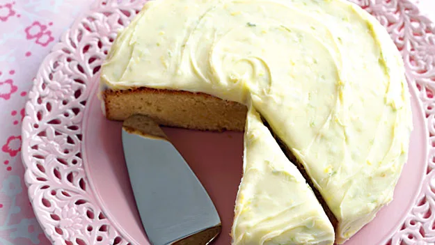 A lemon cake with white icing on a pink lace plate, one slice cut with a cake server beside it.