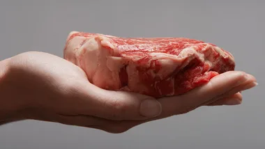 A hand holding a raw, marbled steak against a gray background.