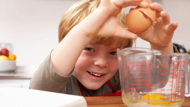 Child cracking an egg into a measuring cup, smiling in a kitchen setting.