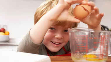 Child cracking an egg into a measuring cup, smiling in a kitchen setting.