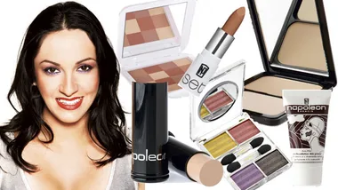 Smiling woman with dark hair surrounded by various colorful makeup products, including lipstick and palettes.