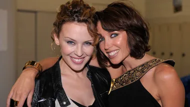 Two women smiling and posing together, one wearing a leather jacket and the other a black dress with a gold embellishment.
