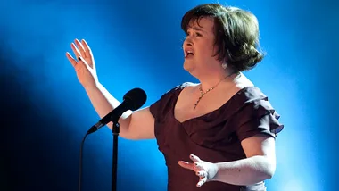 Susan Boyle singing passionately on stage with a microphone in a spotlight.