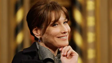 A woman with brown hair smiling, resting her chin on her hand, wearing a dark coat and earrings, sitting indoors.